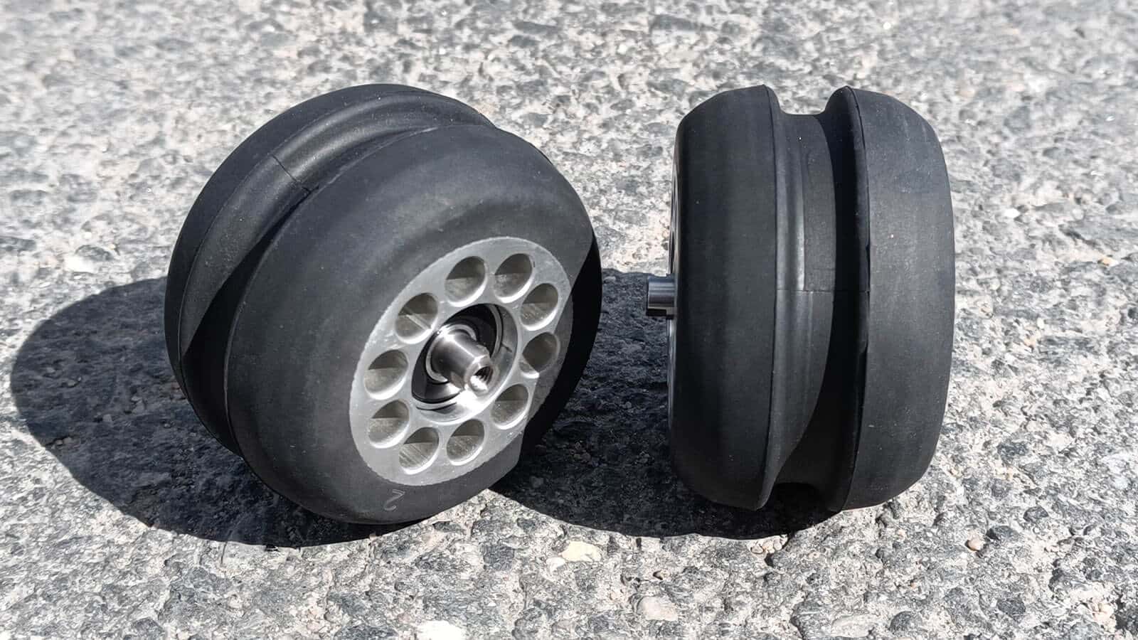 Close-up of two black V-Track wheels with silver hubs, showing the tread and design, placed on a gravel surface.